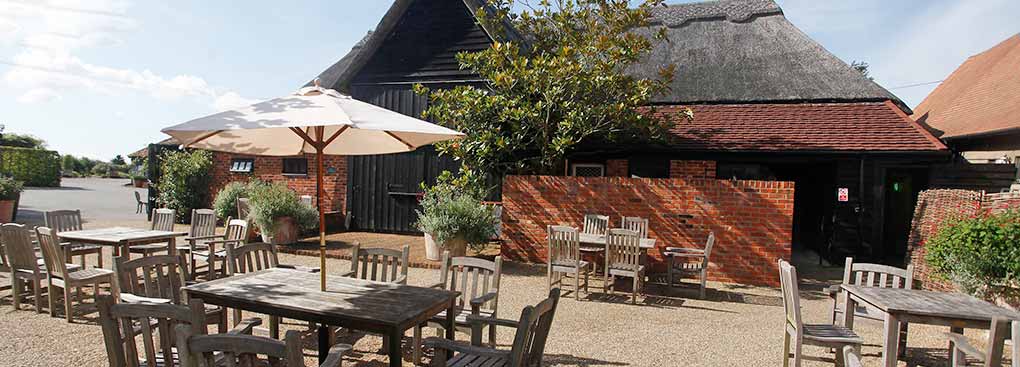 Sunlight illuminates a courtyard full of wooden tables and parasols outside a large thatched barn