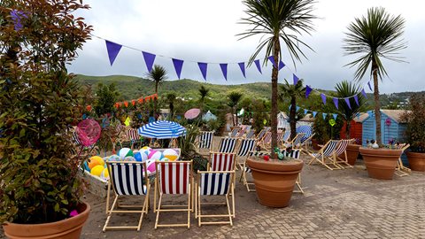 Colourful huts surround a beach set against the backdrop of the Malvern Hills. Potted palm trees, coastal grasses and deckchairs complete the scene