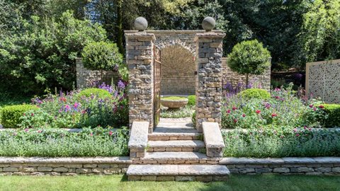 This garden features meticulously crafted drystone walling and dark pink and deep purple roses with perennials in shades of purple, burgundy, pink and blue.