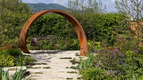 A Corten steel arch sits among rich, wildlife-friendly beds of tangerine <i>Geum</i>, <i>Astrantia</i> and vibrant <i>Euphorbia</i>, perfectly blending new materials with natural planting