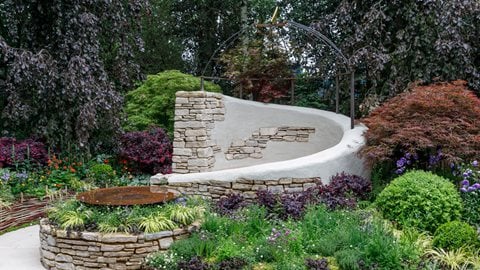 A stone and partially-rendered wall curves around a central seating area that is surrounded with a purple planting scheme and woven willow borders filled with edible plants and herbs