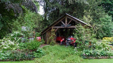 A vintage car sits in a garage that is being slowly reclaimed by nature, with red rambling roses, grasses and orange geums gradually taking over the space
