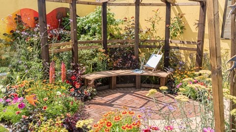 A richly planted garden illustrating the work of the charity, Start in Salford. It features a central seating and patio area, pergola and a profusion of brightly-coloured flowers and foliage