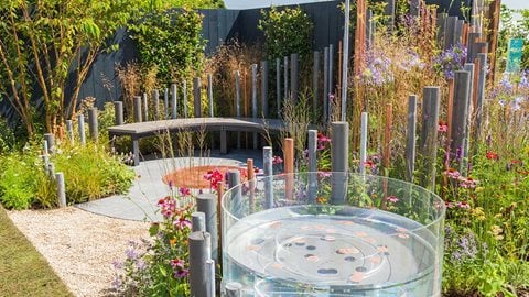 A contemporary glass wishing well sits in the foreground amongst vibrant colourful planting, evoking the feeling of hope for families