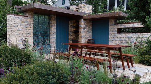 A stone-built shelter, complete with a green roof, overlooks an array of purple planting that includes alliums, purple verbascum and cirsium as well as an aged pear tree in a meadow