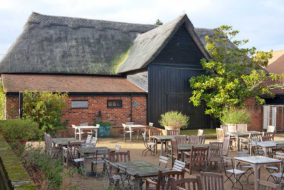 Empty tables and chairs are seen in a sunlit courtyard in front of a large barn with a thatched roof